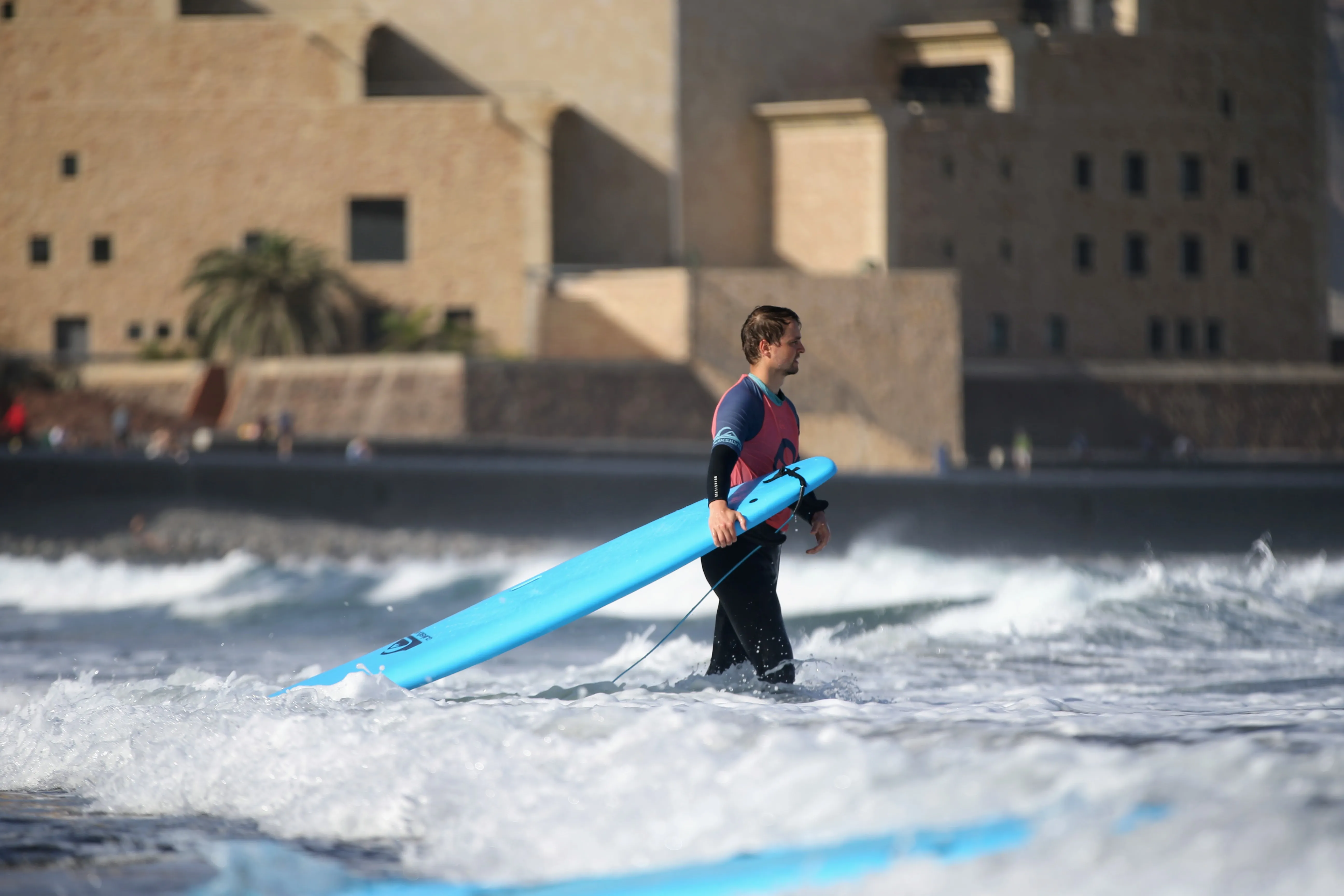 Connor walks into the shallow waves holding his turquoise surfboard on his right side. A large sandstone building rises in the background
