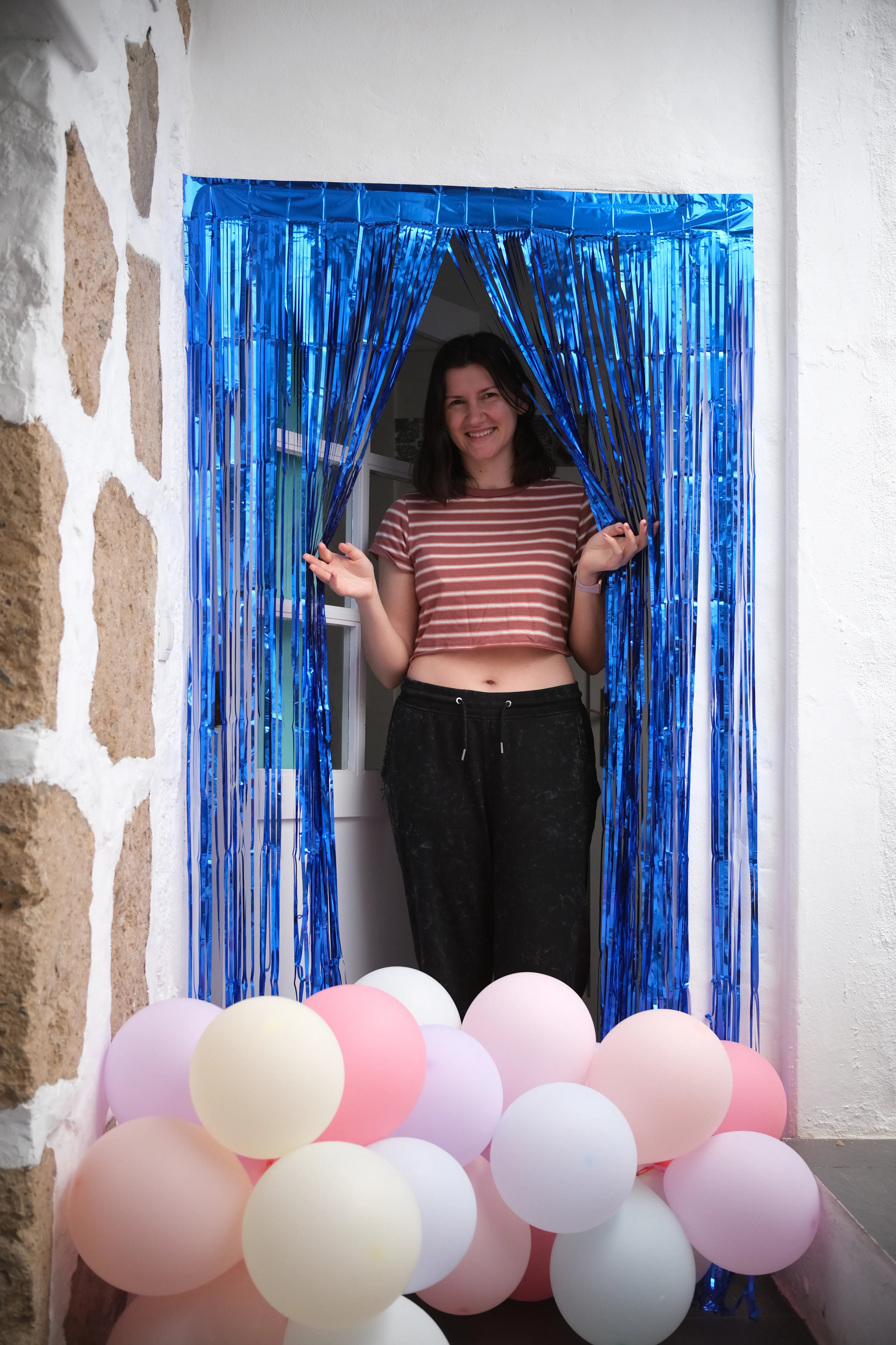 Jillian peeks through a door curtain of shimmering blue ribbons. The doorway is clogged by clusters of pastel colored balloons.
