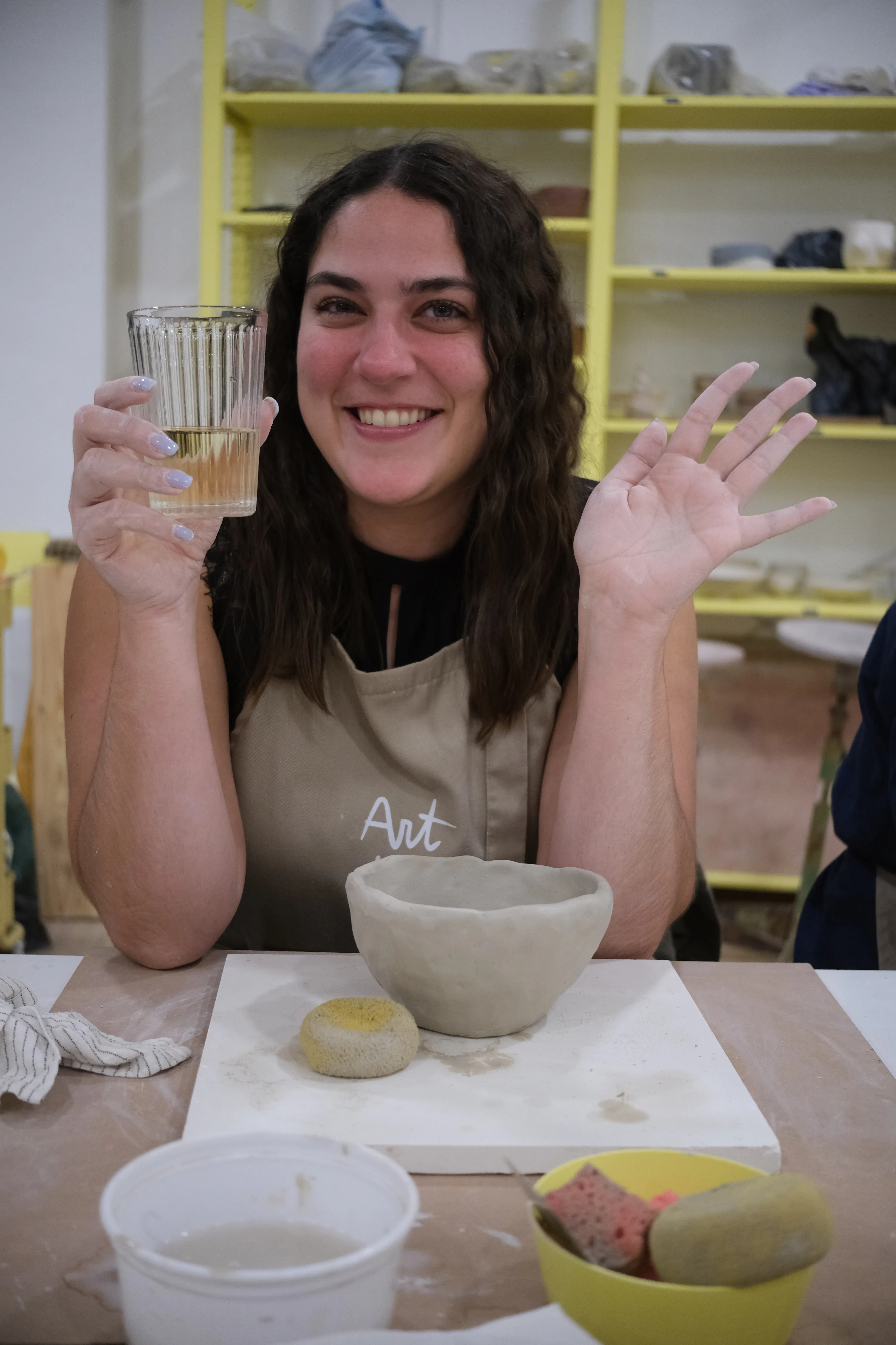 Alessandra smiles and waves at the camera while holding a glass of wine in her other hand. A bowl of clay and some sponges lie on the table in front of her.