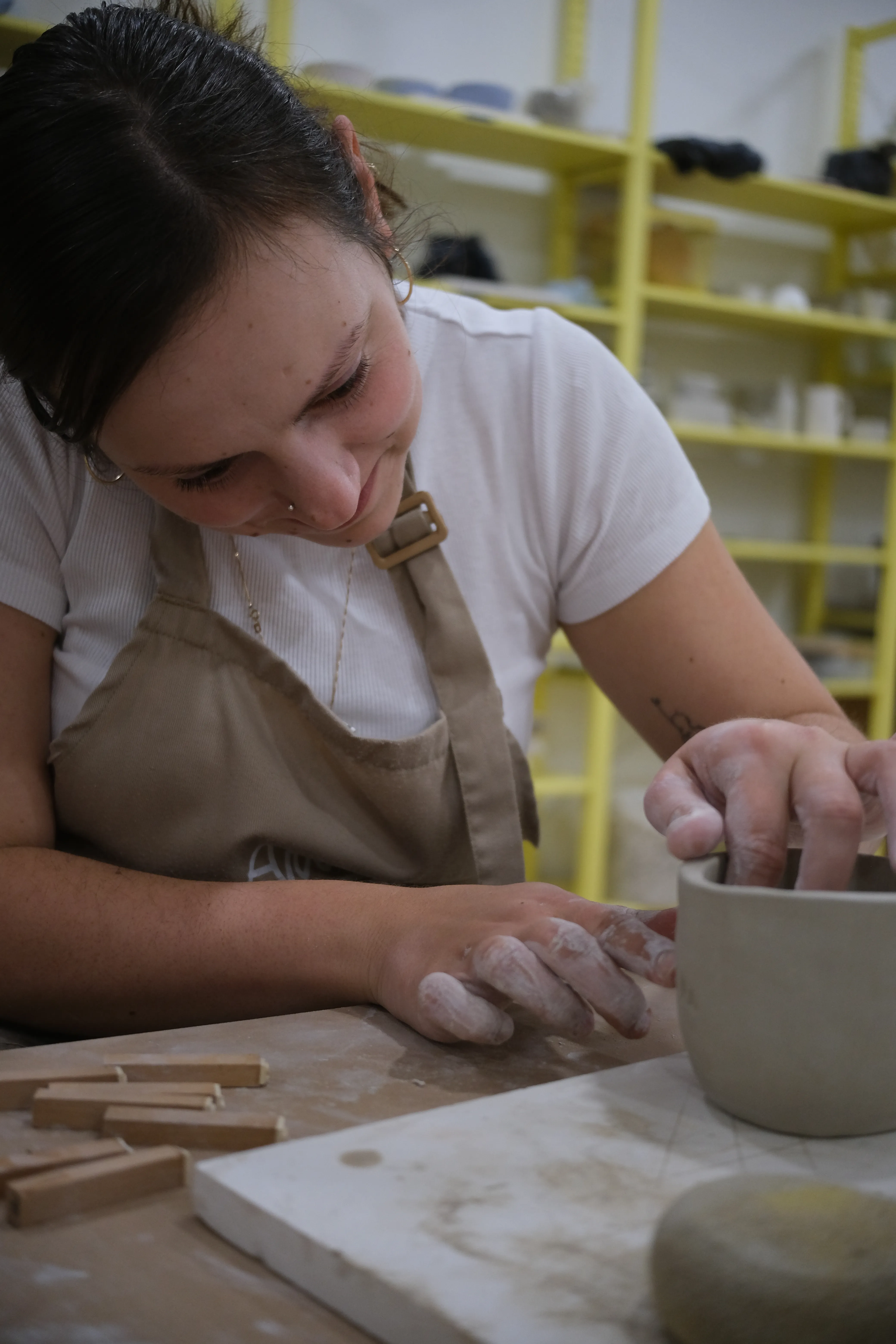 Ena focuses on carefully shaping a bowl of clay. Her fingers are covered in a thin layer of white, dry clay.