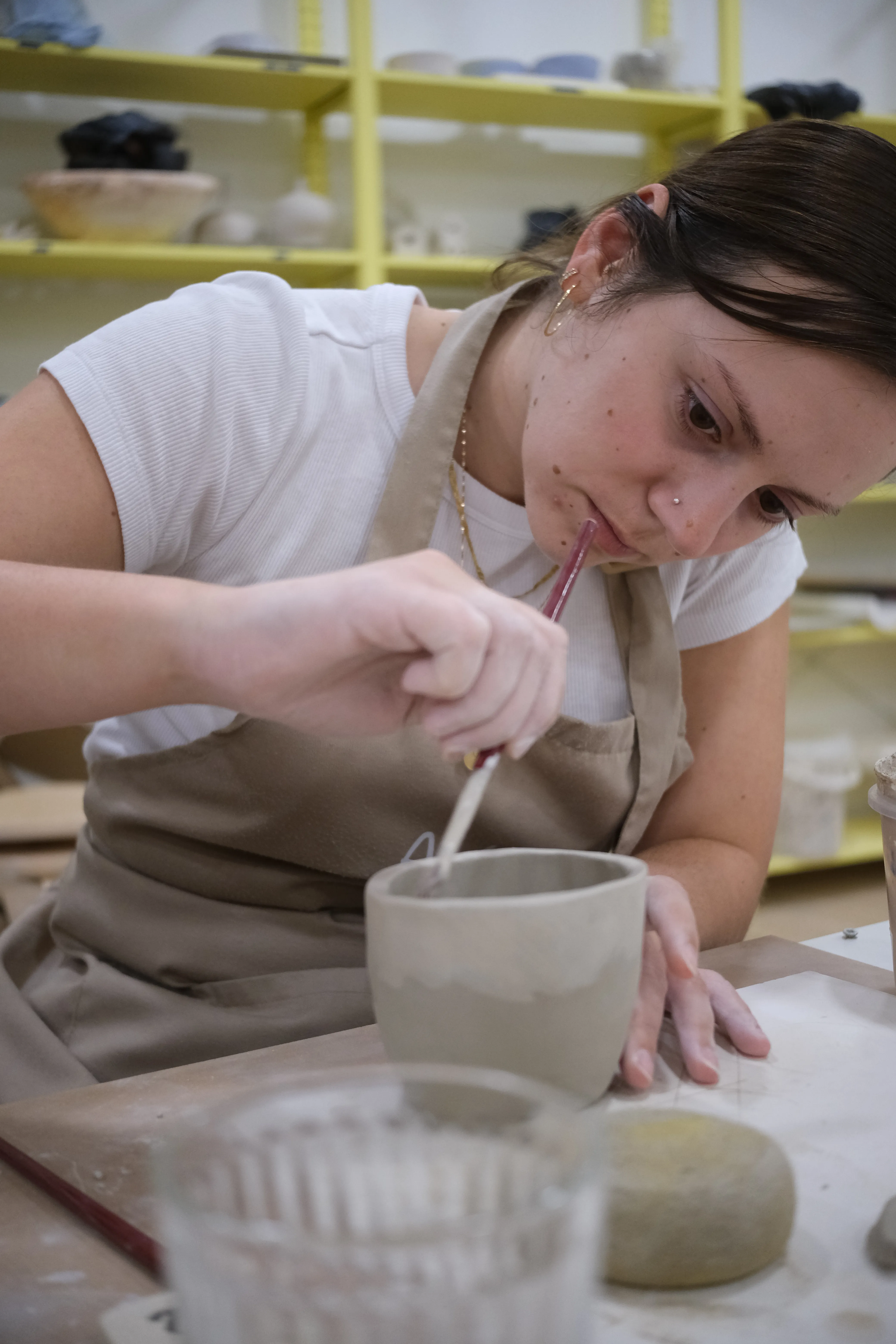 Closeup of Ena painting the outside of her cup an off-white color. She is wearing a white t-shirt and ocher apron.