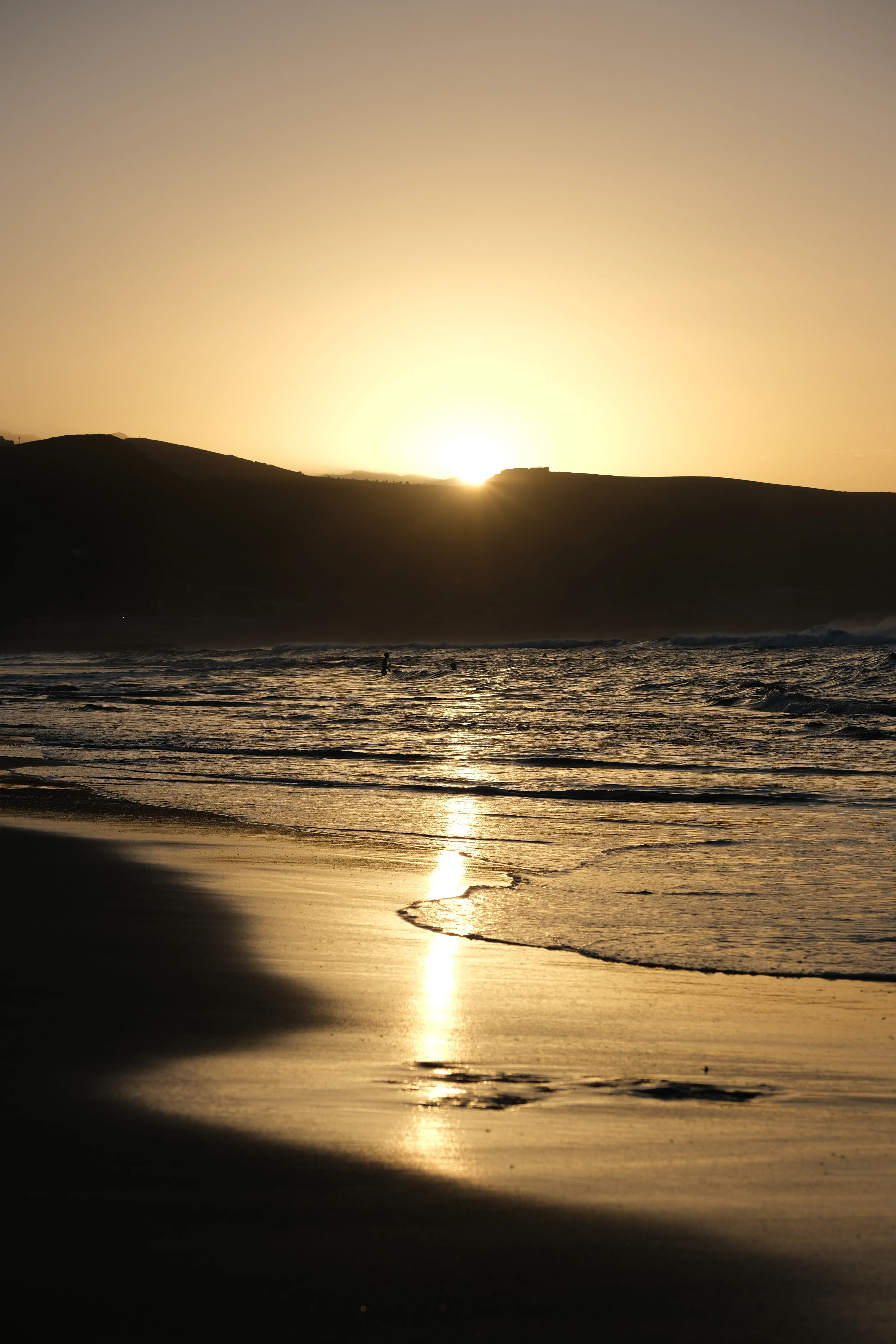 Ocean waves gently wash onto the beach in the golden-yellow light of the sun setting behind the hills of Gran Canaria.
