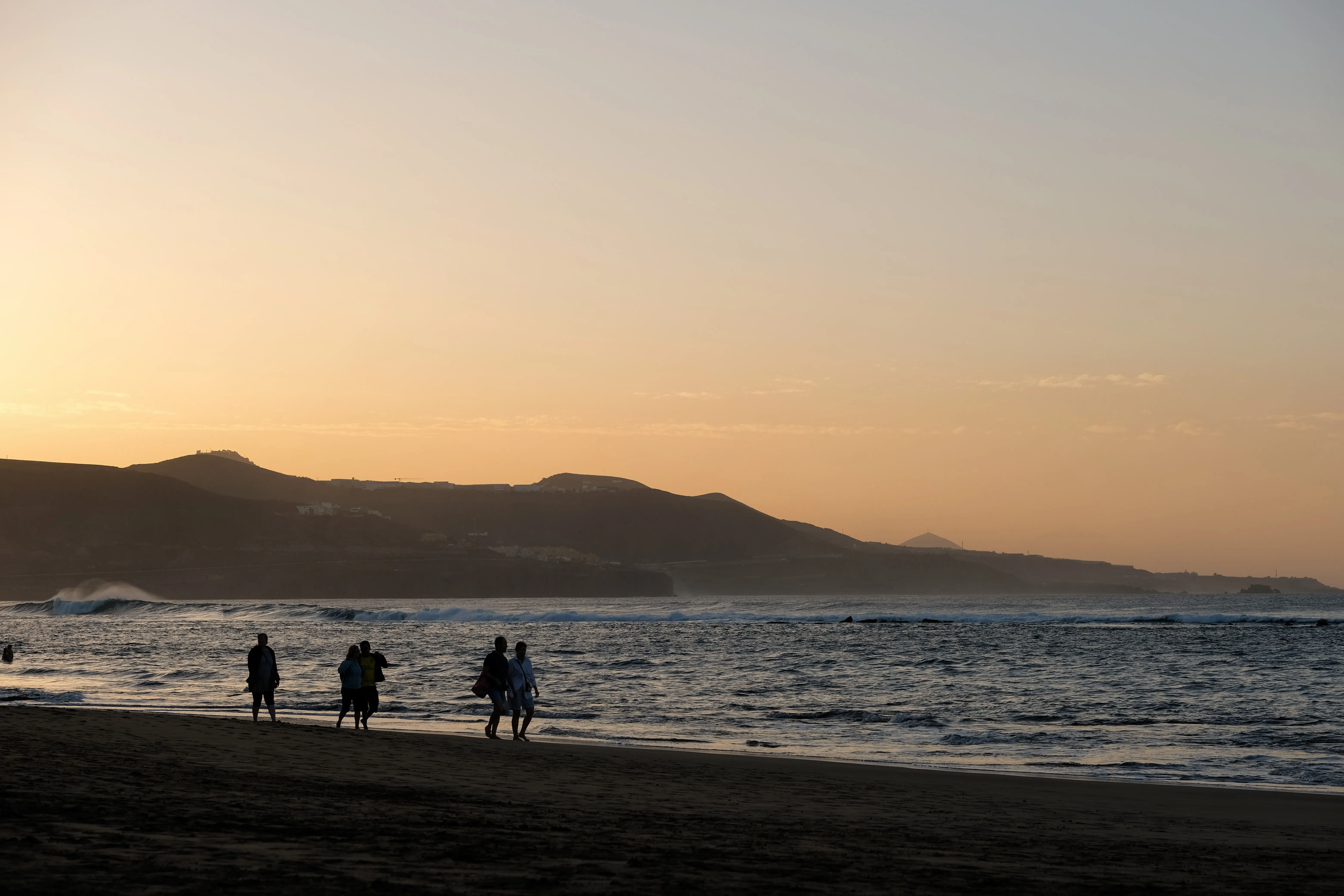 Silhouettes of people walk on the beach at golden hour. The setting sun bathes the sky and calm waves in an orange light.