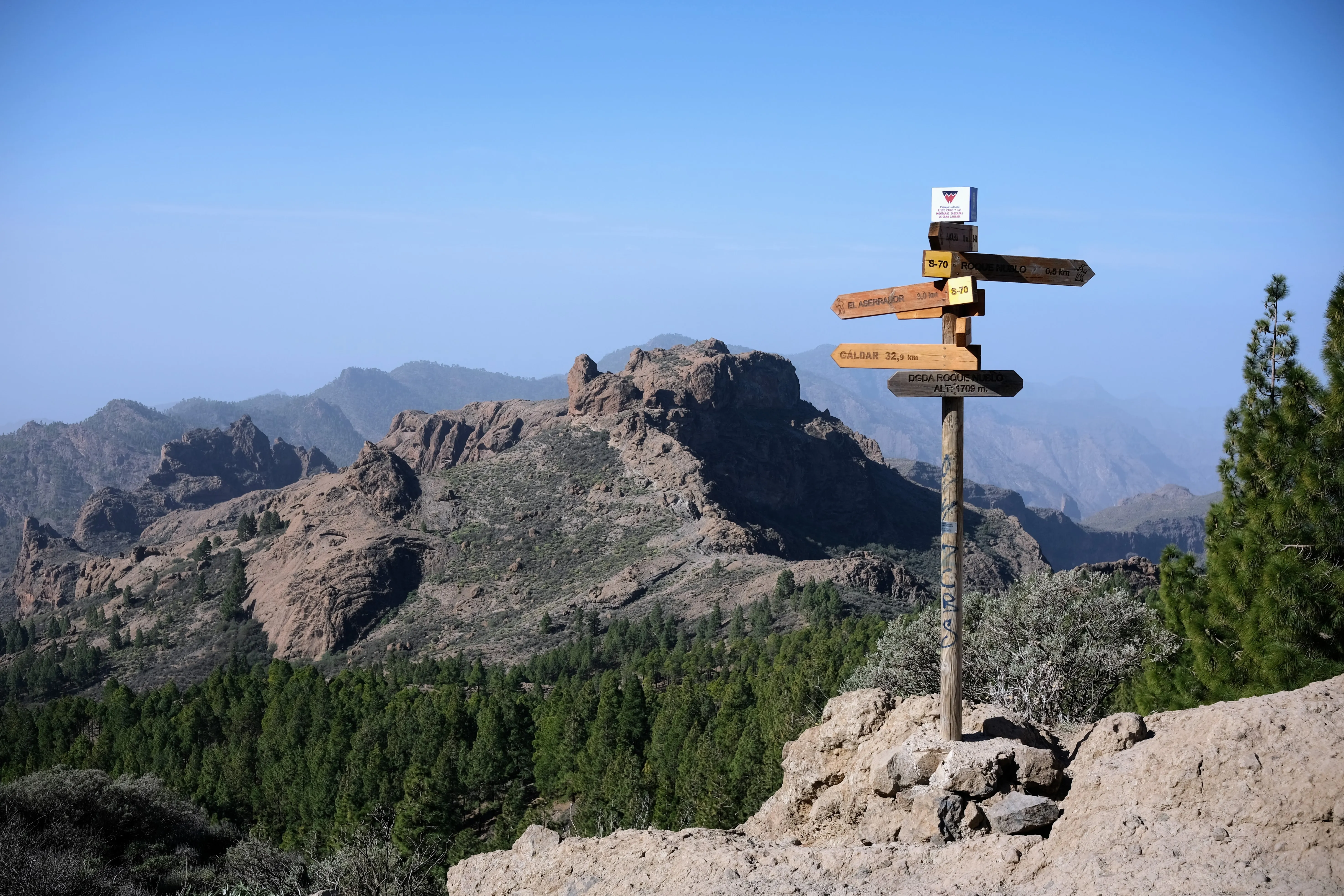 Panoramic view of a rocky hill surrounded by a deep green pine forest. In the foreground, a wooden sign points the way against the backdrop of the hazy blue sky.