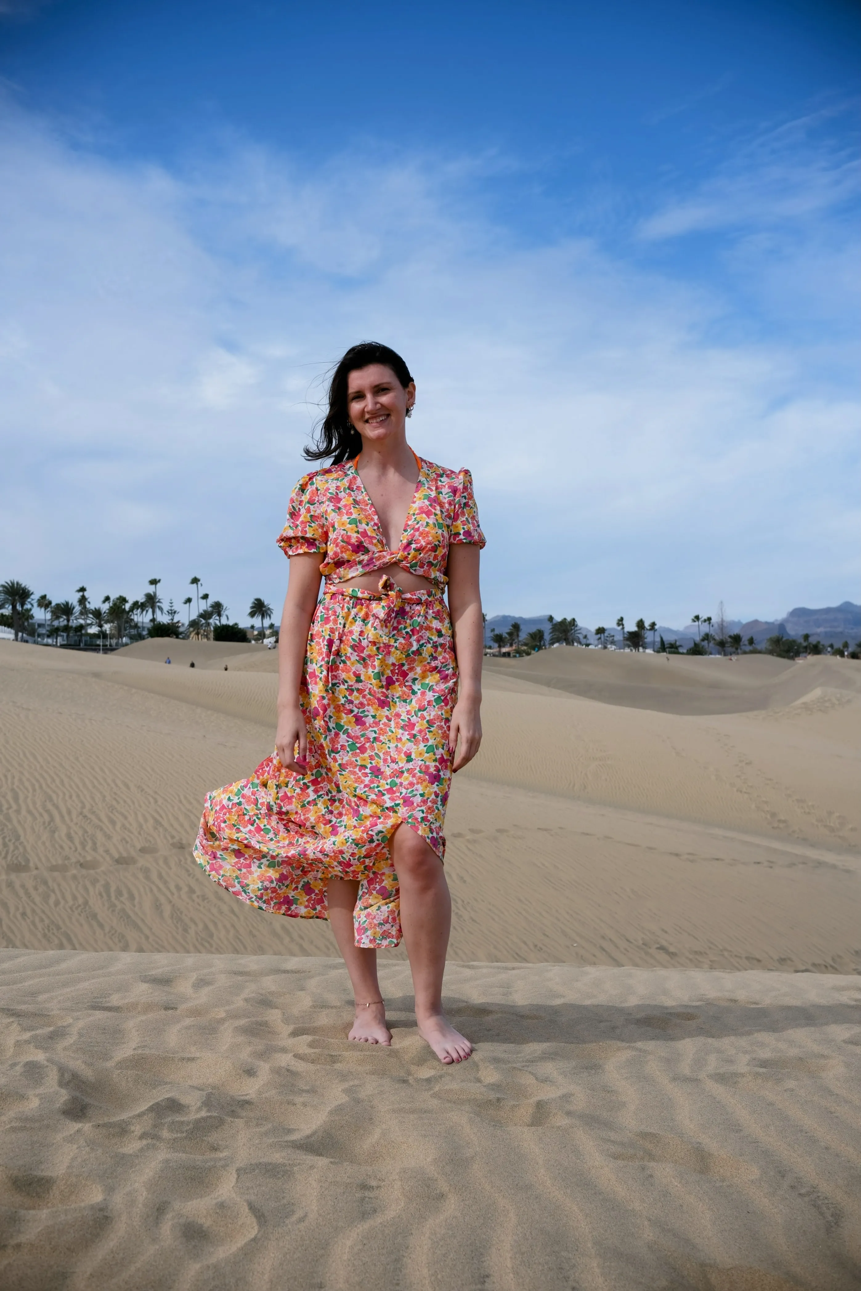 Jillian in a colorful summer dress stands barefoot in the sand dunes of Maspalomas.