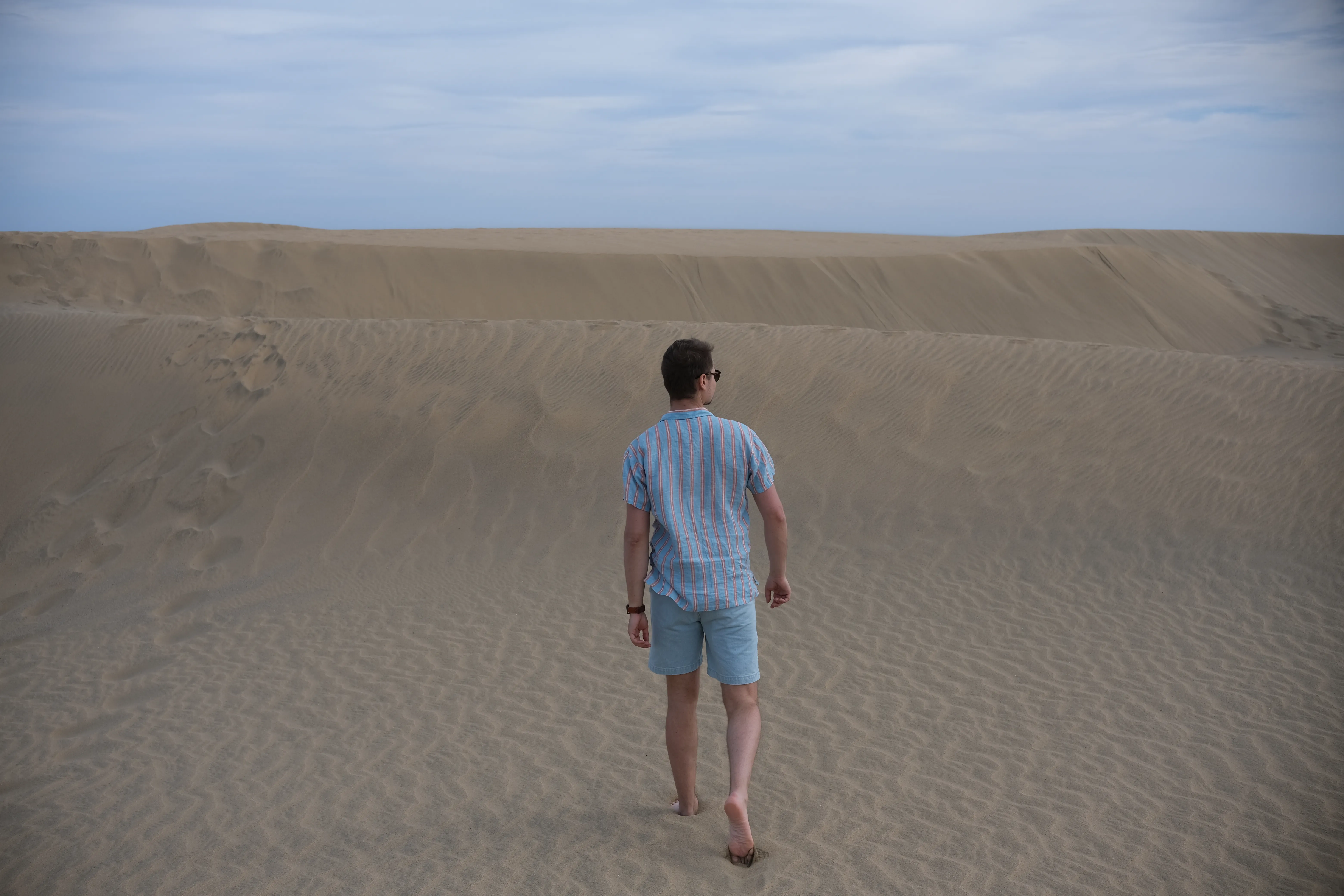Connor walks barefoot towards a rolling sand dune. He is wearing a light blue shirt and denim shorts.