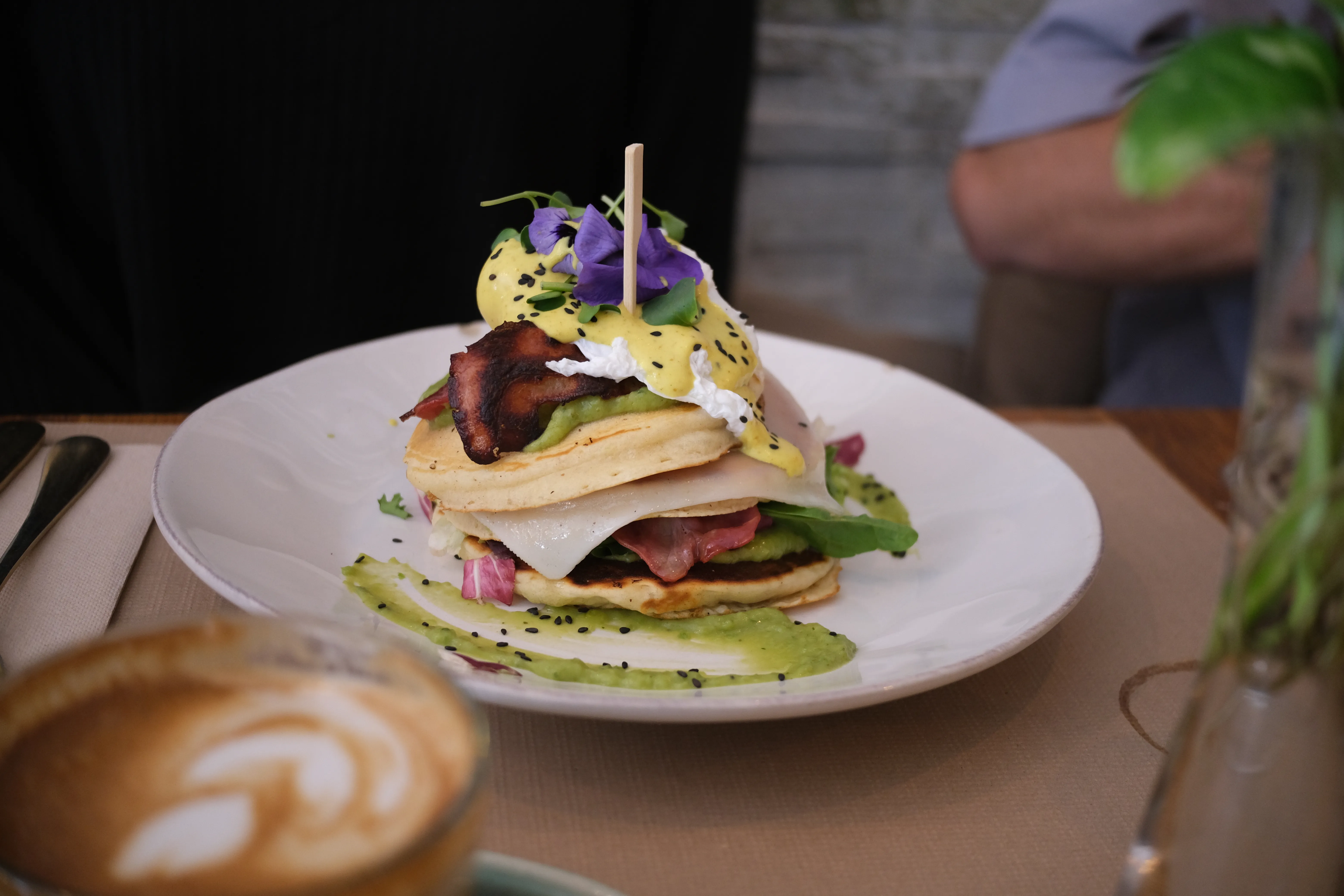 A stack off pancakes interlaced with bacon, eggs, cheese and avocado, artfully decorated with edible flowers and black sesame seeds.
