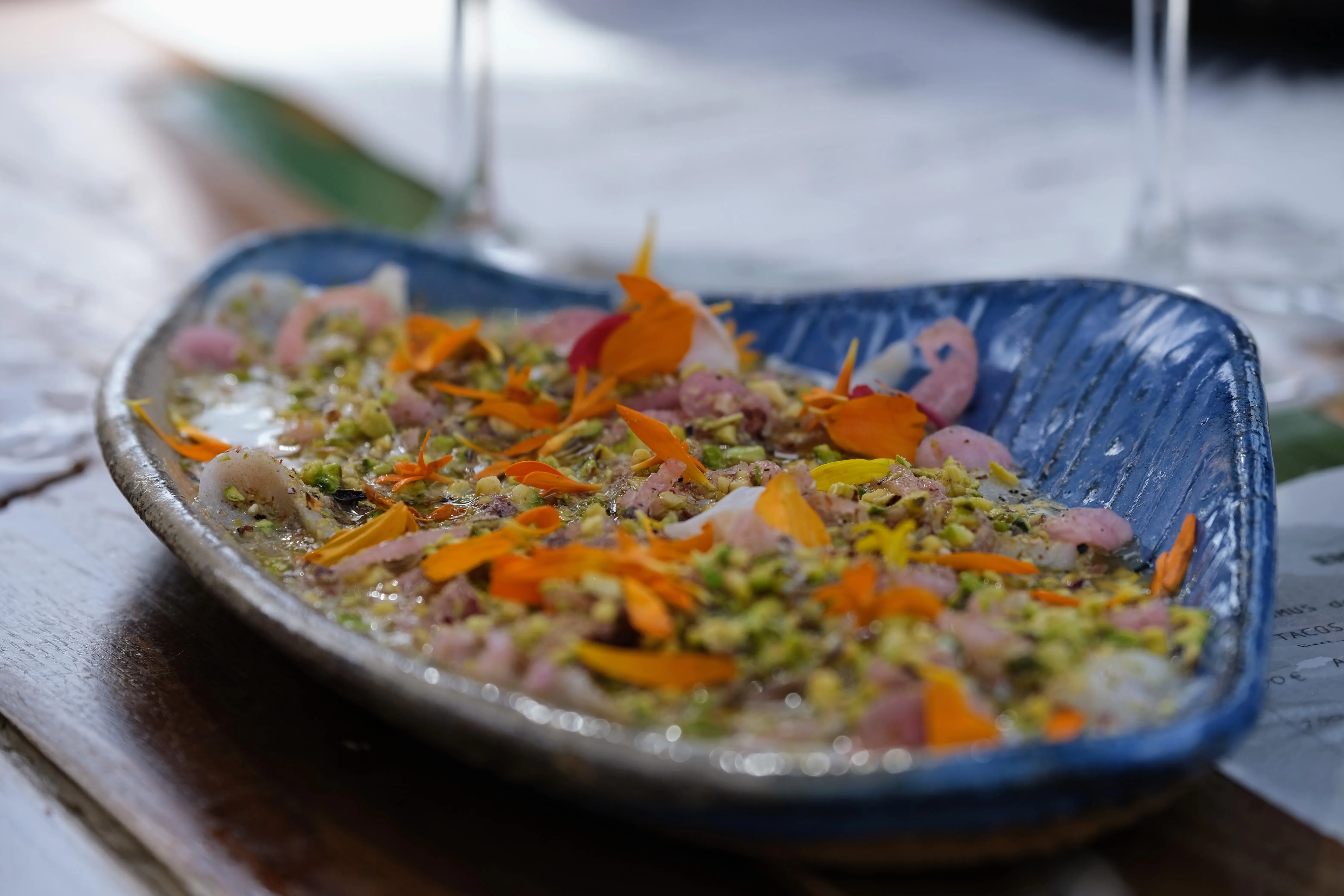 A blue handmade plate of cod ceviche with chopped pistachios and orange edible flower petals.