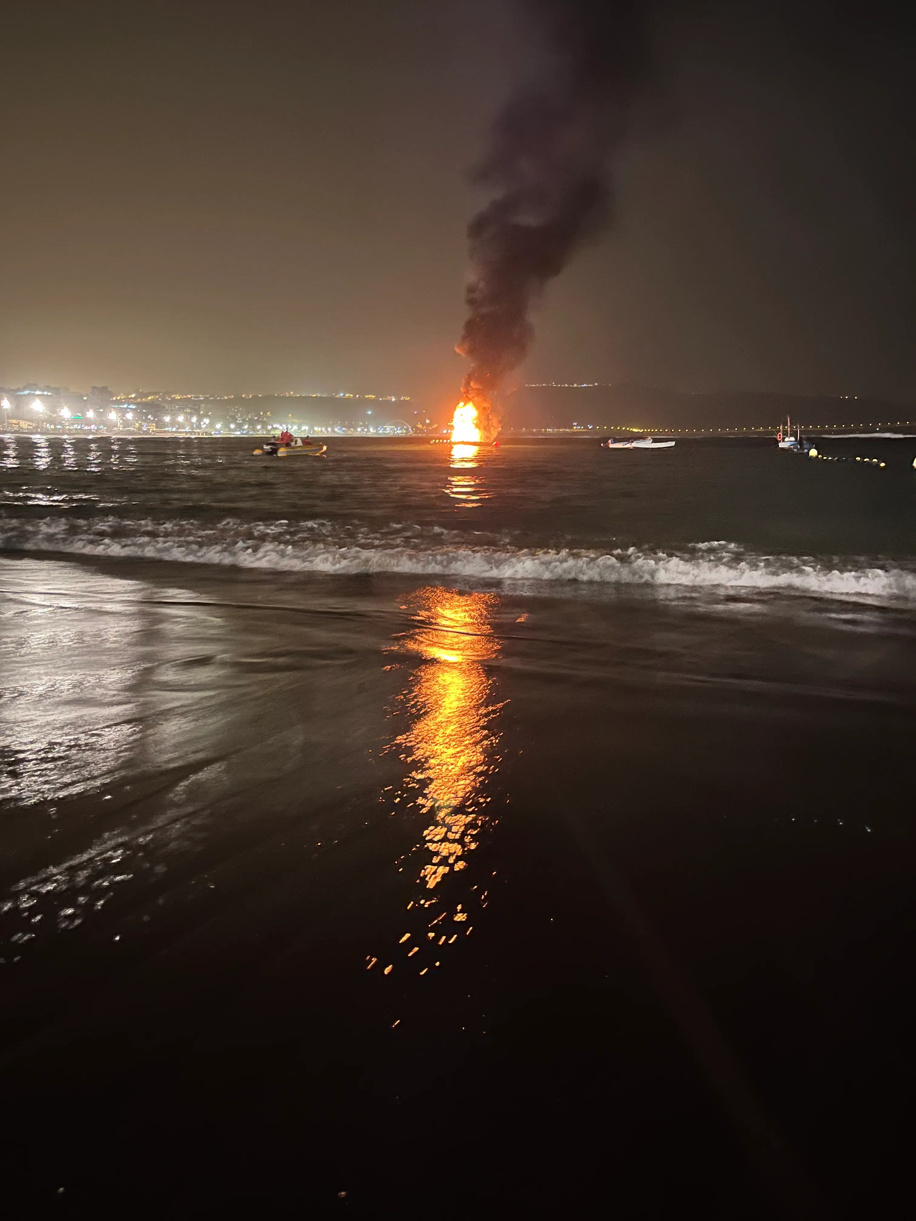 A bright yellow flame atop a barge spews black plumes of smoke into the night sky, illuminating the surrounding ocean waves.