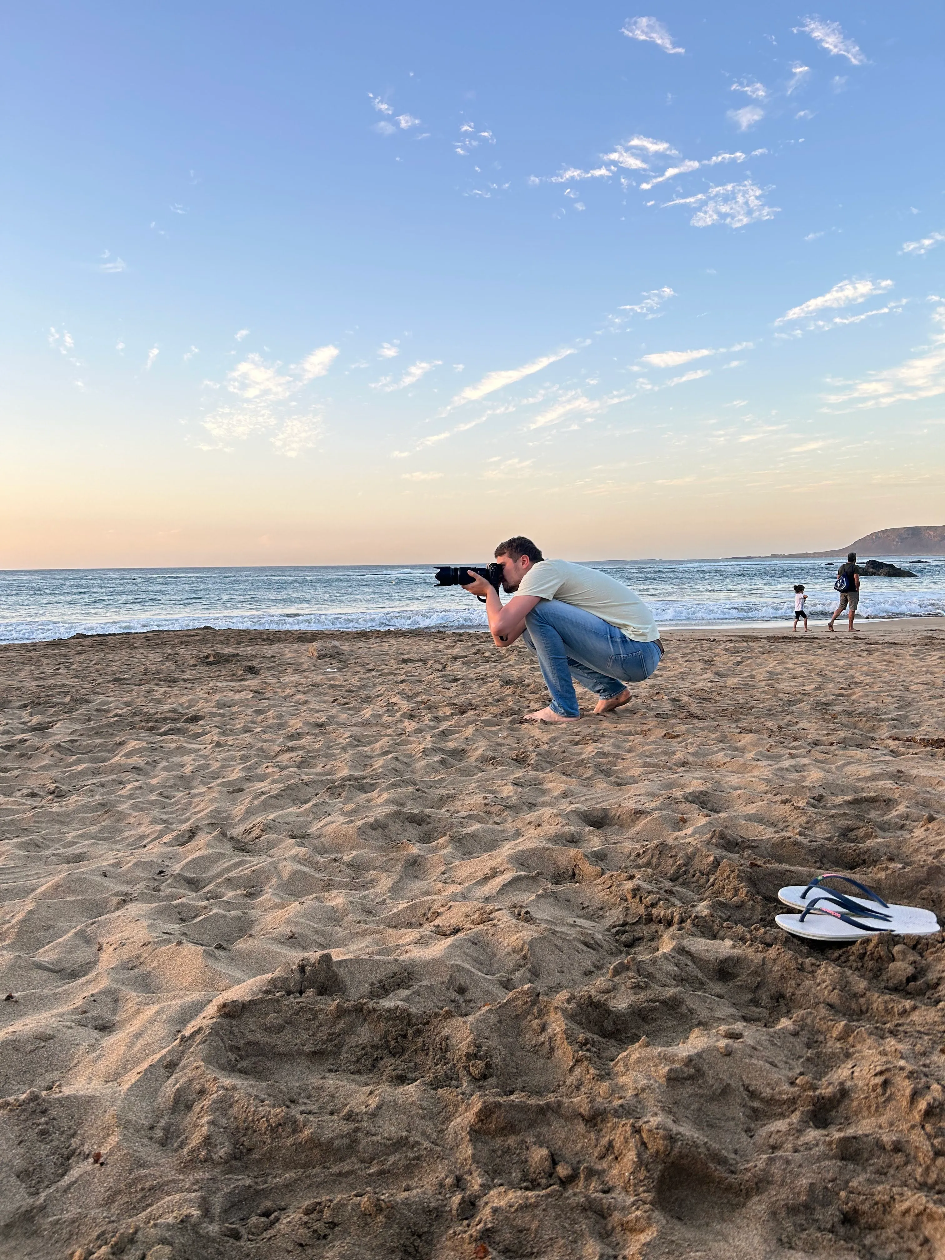 Connor crouches in the beige sand. He is looking through a camera with a long lens, focused on taking a picture of the sunset in the distance.