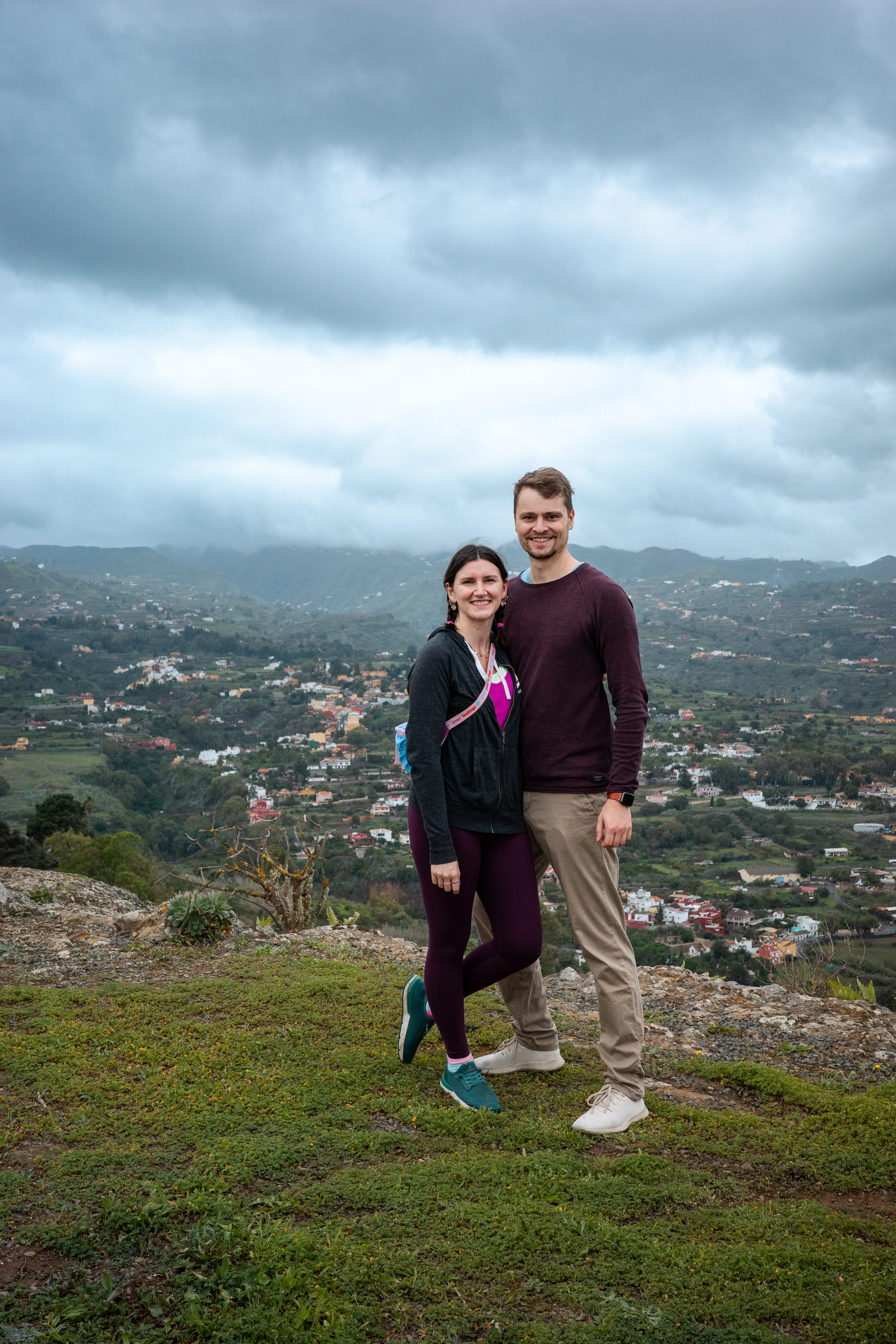 Jillian and Connor stand arm in arm on a green meadow that drops off into the valley behind them. The sky is covered in grey clouds.